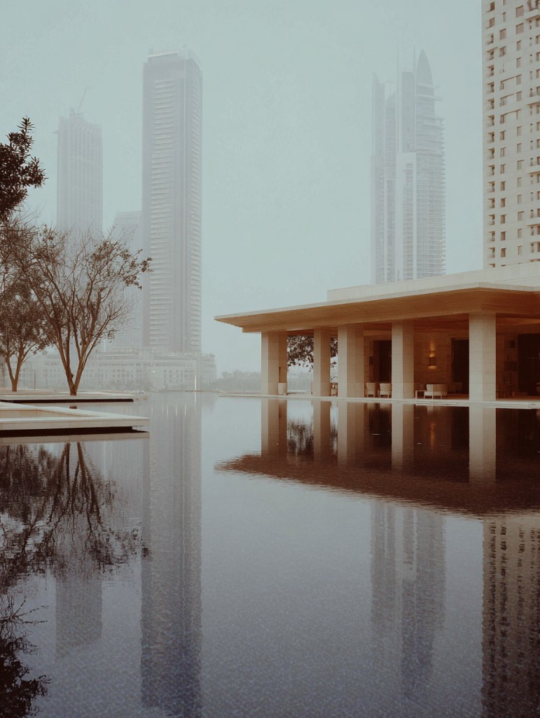 A long mirror pool reflecting modern towers under a pale, hazy sky